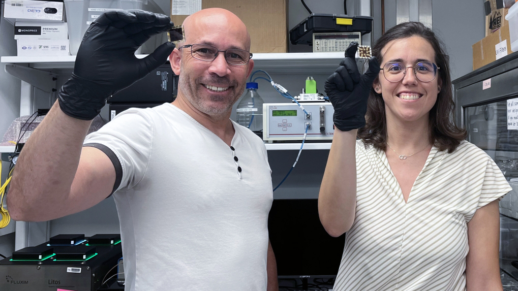 Two faculty members in a laboratory hold up small electronic components while wearing black gloves.